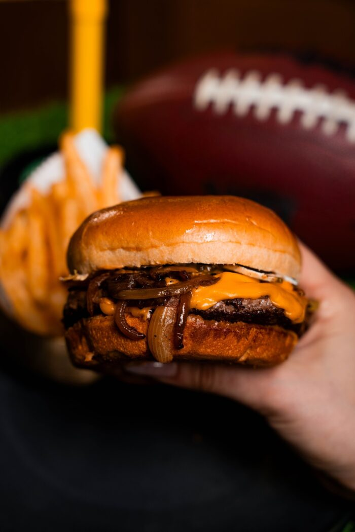 Where To Enjoy the Best Burger in Salt Lake City Close-up of a cheeseburger with grilled onions held in front of a football and fries.