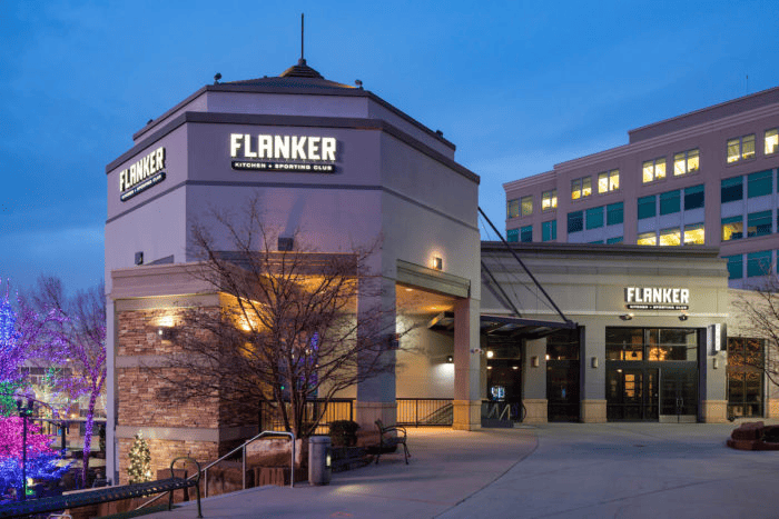 Exterior view of Flanker Kitchen + Sporting Club at dusk with illuminated signs and nearby festive lighting.
