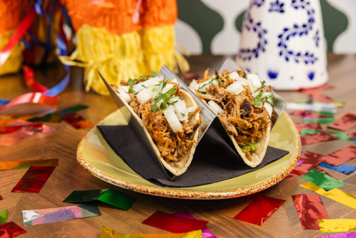 Two shredded meat tacos garnished with chopped onions and cilantro, served on a plate with festive confetti and decorations.