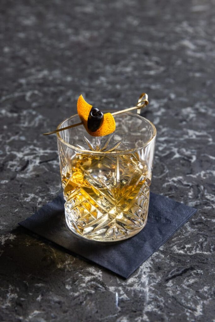 Crystal glass of whiskey on a dark napkin, garnished with an orange twist and cherry on a cocktail pick.