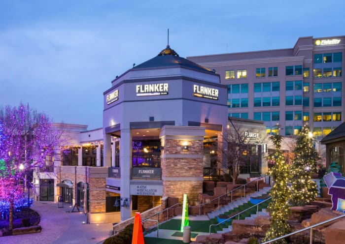 A multi-level building with the name "FLANKER" illuminated on its sides at dusk. The building is surrounded by decorated trees with white and colorful lights, and a miniature golf course with stairs leading up to the entrance. In the background, a larger building with a Fidelity sign is visible.