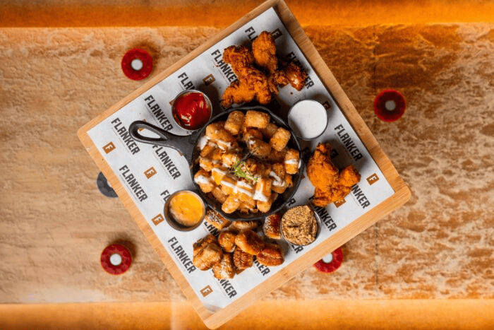 Overhead view of a tray with fried chicken, tater tots, dipping sauces, and other appetizers served on Flanker-branded paper.