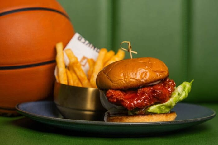 Explore Christmas Bars in Salt Lake City Crispy chicken sandwich with lettuce on a toasted bun, served with golden French fries in a small bowl, placed beside a basketball on a green booth backdrop.