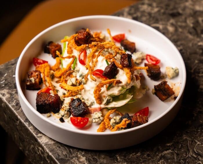 Explore Christmas Bars in Salt Lake City Wedge salad served on a white plate with crispy bacon, cherry tomatoes, crumbled blue cheese, and fried onions, placed on a dark marble table