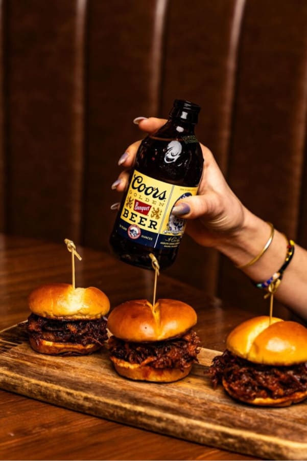 Explore Christmas Bars in Salt Lake City A hand holding a Coors Banquet beer bottle above a wooden board with three pulled-pork sliders.