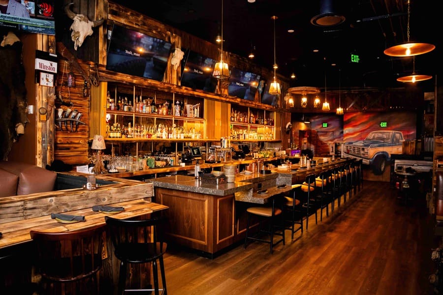 Rustic sports bar interior with a long wooden bar, liquor shelves, mounted TVs, and warm lighting.