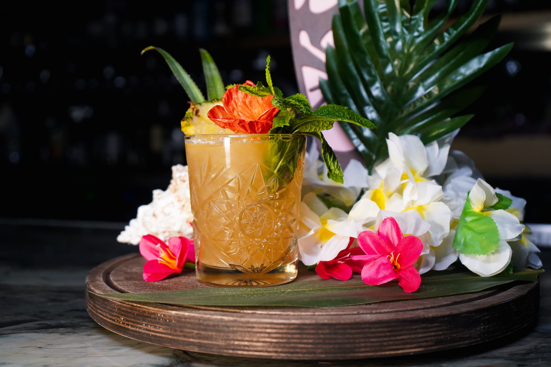 Tropical My Tai cocktail with pineapple garnish, mint, and tropical flowers on a wooden serving tray.