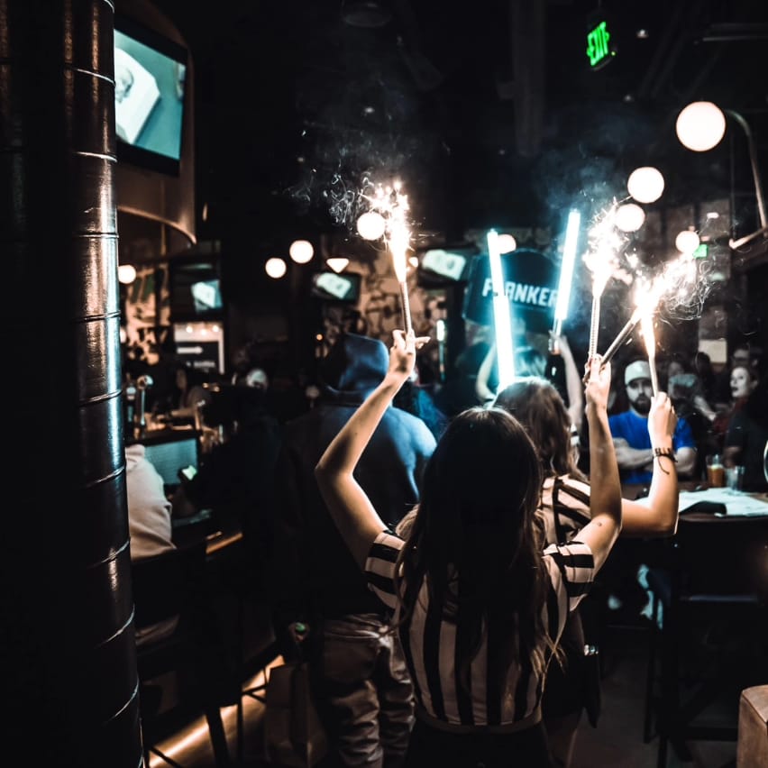 Flanker staff carrying lit sparklers through a lively, dimly lit bar during bottle service.