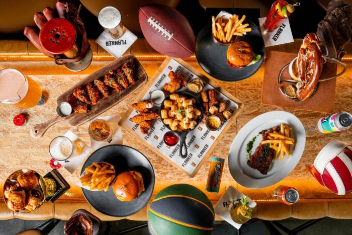 Image pop-up Overhead view of burgers, fries, wings, chicken tenders, steak, pretzel, drinks, and sports-themed decor arranged on a table.