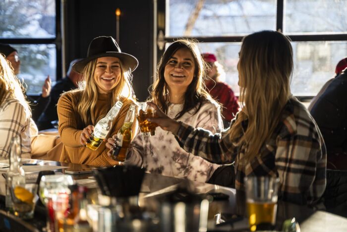 Image pop-up Three guests smiling and raising drinks together while seated at a sunlit sports bar.