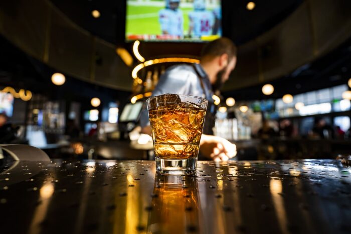 Image pop-up Glass of whiskey with ice on a bar counter with a bartender and sports screens in the background.