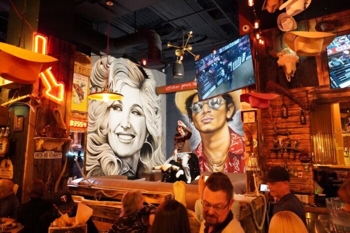 Image pop-up Western-themed bar with a mechanical bull, colorful murals, cowboy decor, and televisions.