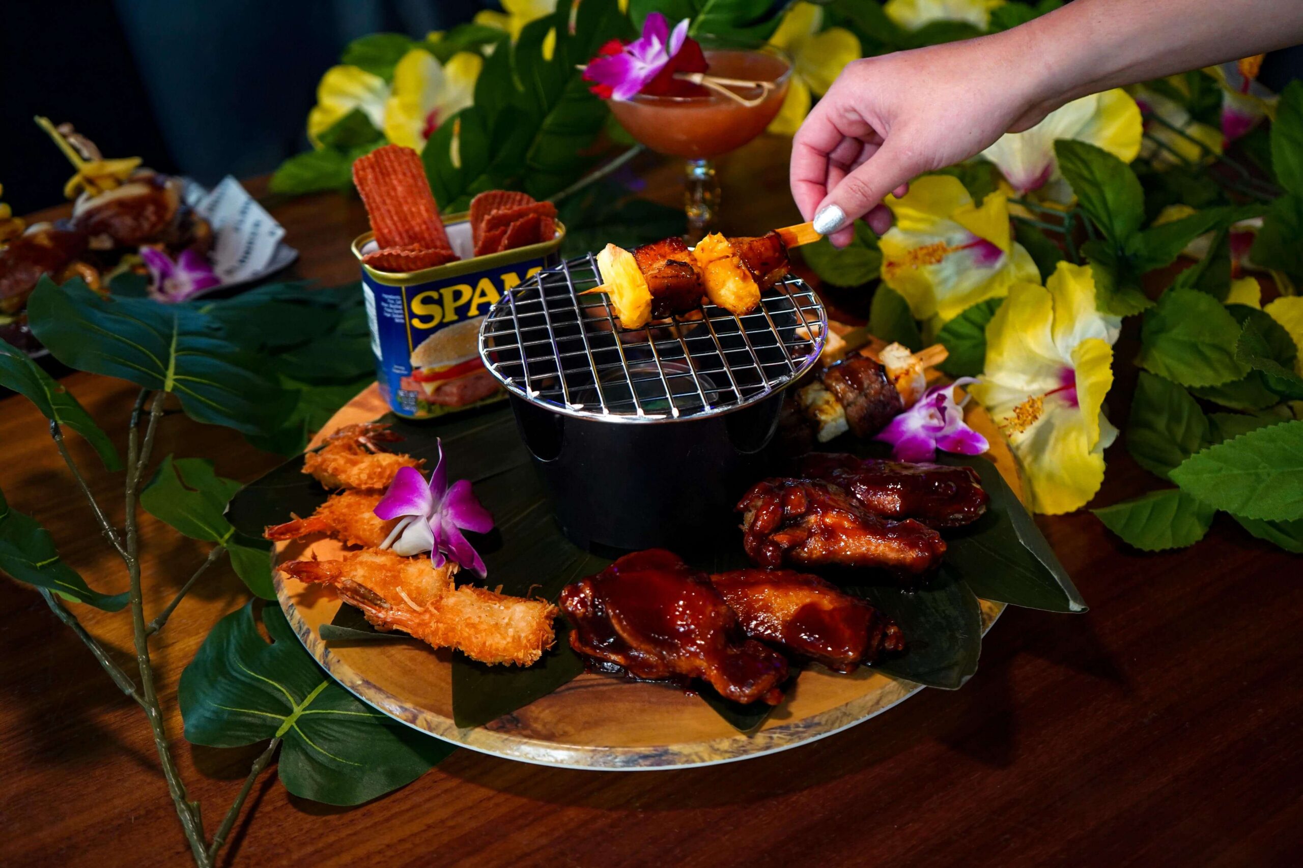 Tropical food platter with glazed wings, fried shrimp, grilled skewers, and crispy snacks surrounded by colorful flowers and greenery.