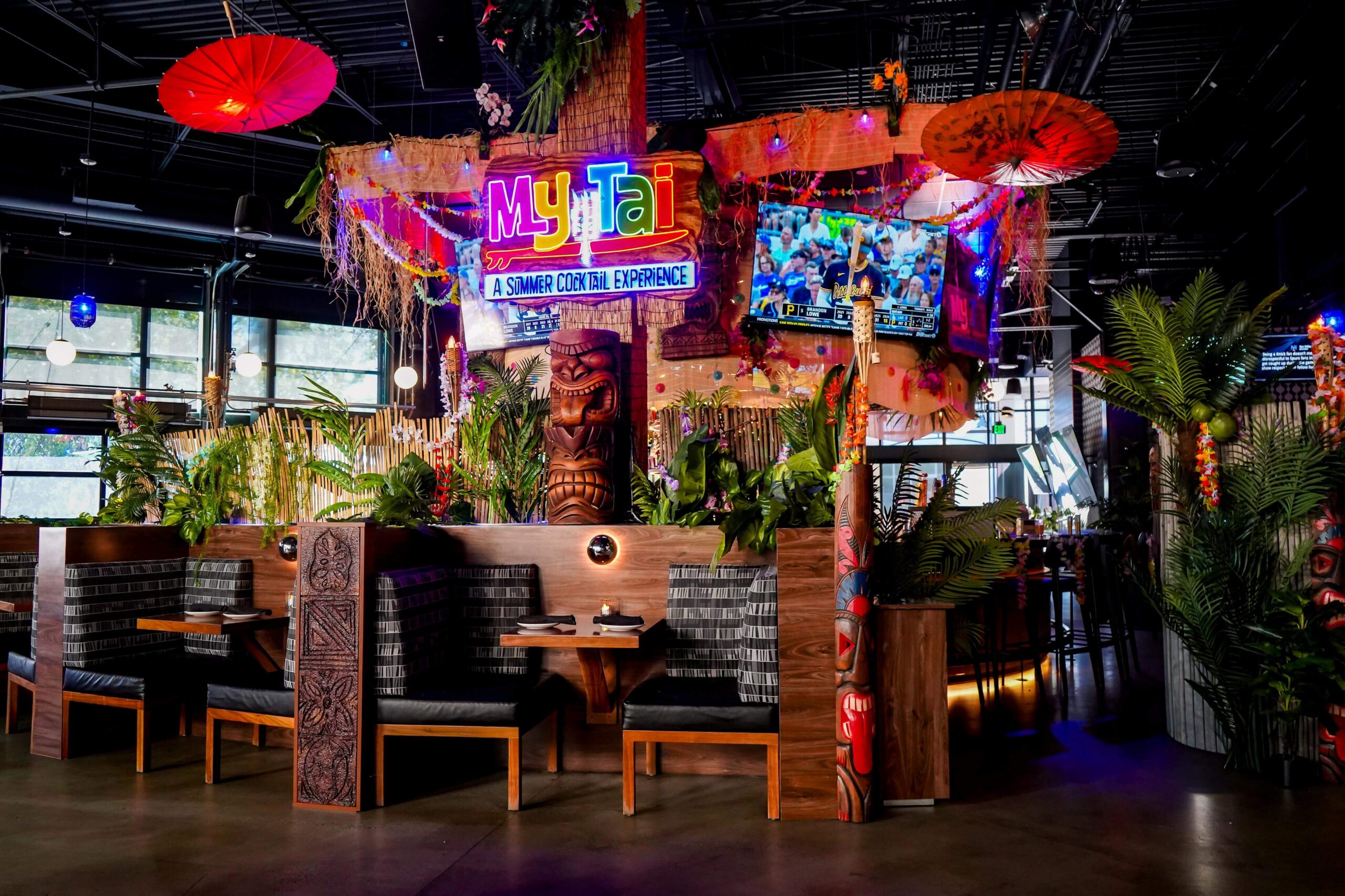 Tropical-themed My Tai dining area with booth seating, lush greenery, colorful lighting, tiki décor, and red parasols.